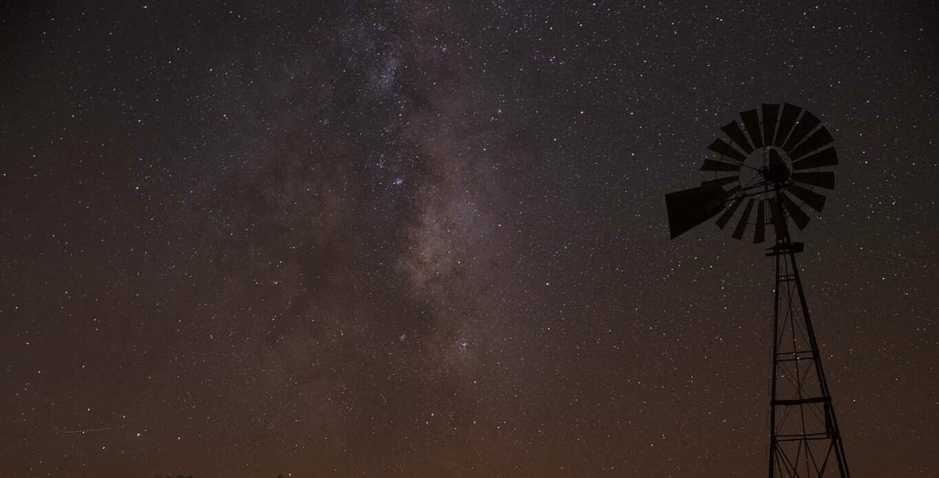 Silhouette of a traditional ranch windmill standing beneath a star-filled night sky. The Milky Way stretches across the darkness, creating a dramatic backdrop over the rural landscape.