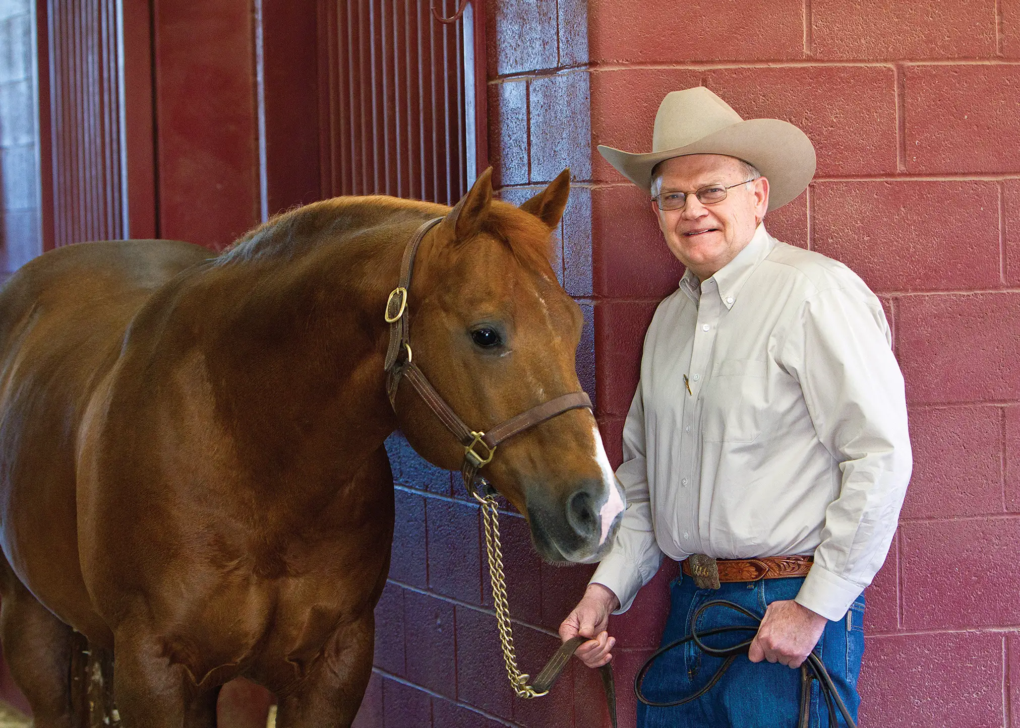 Dr. Glenn Blodgett in a button-up with his glasses and cowboy hat holding the reins of a horse.