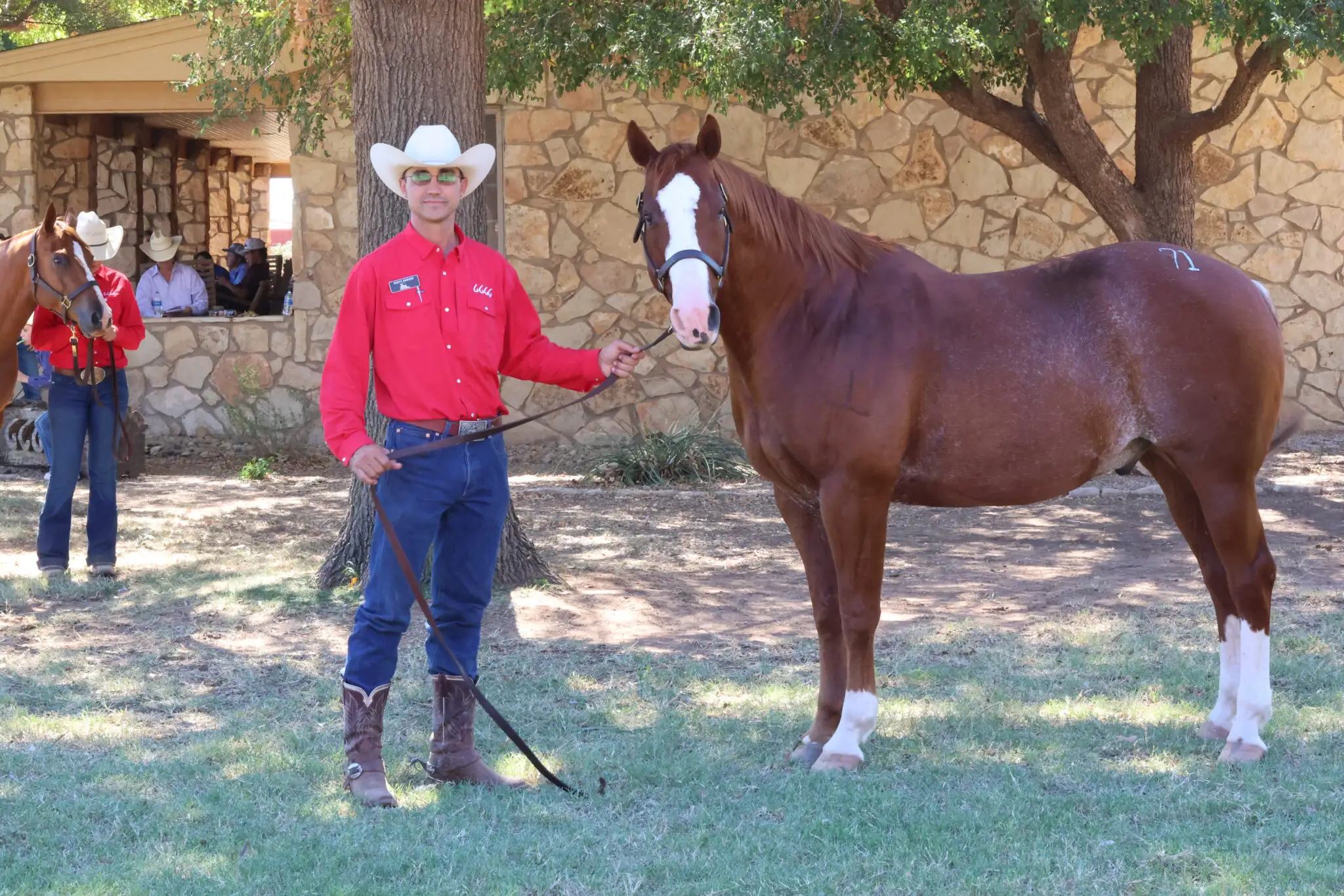 A ranch representative in a red Four Sixes Ranch shirt stands beside a chestnut horse beneath shade trees on the ranch grounds.